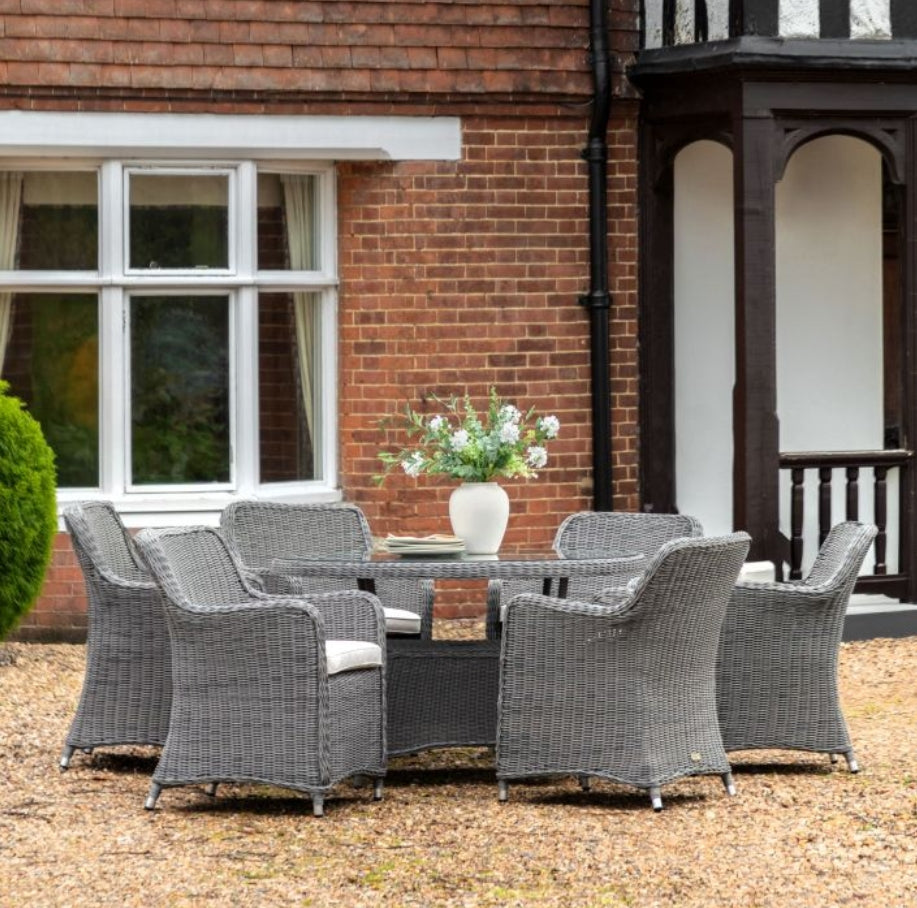 A 7-piece outdoor dining set featuring a round glass-top table and six grey woven wicker armchairs, shown against a plain white background. Available at Albury House Interiors.