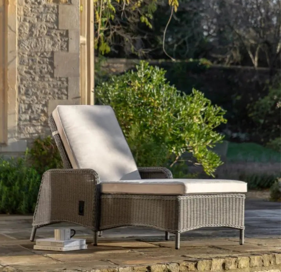 An outdoor reclining wicker Lounger with a tall backrest, featuring light beige cushions on the seat and back, set against a plain white background.