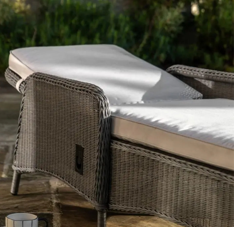 An outdoor reclining wicker Lounger with a tall backrest, featuring light beige cushions on the seat and back, set against a plain white background.