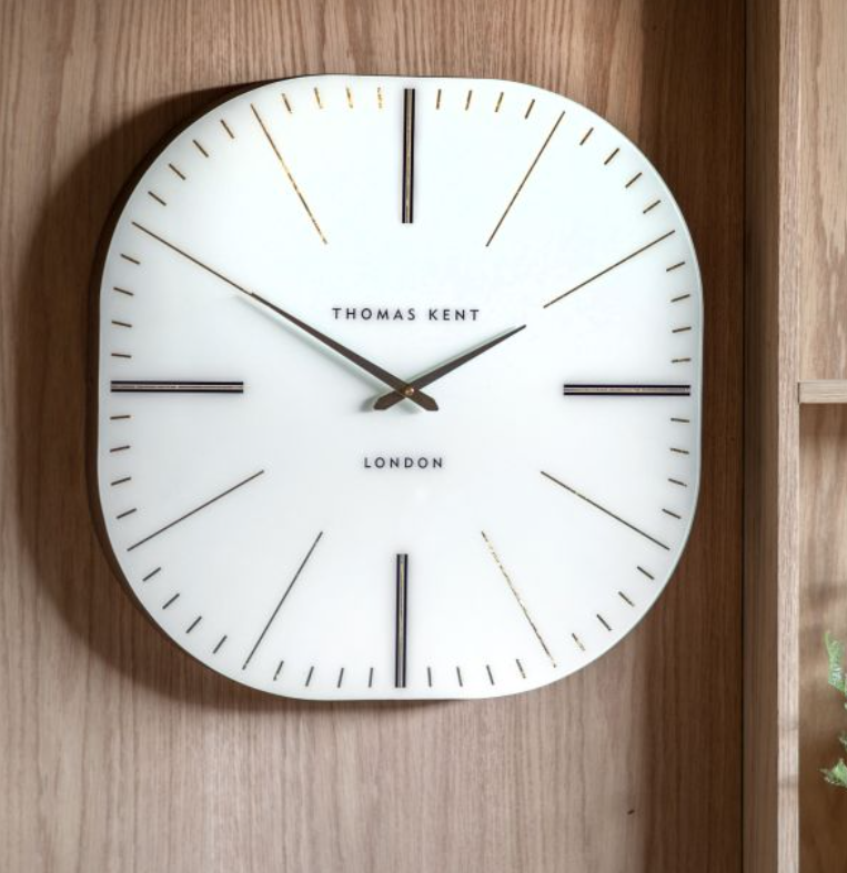 A Thomas Kent London wall clock with a white face, black hands, and minimalist black hour markers, mounted on a warm wood-paneled wall.