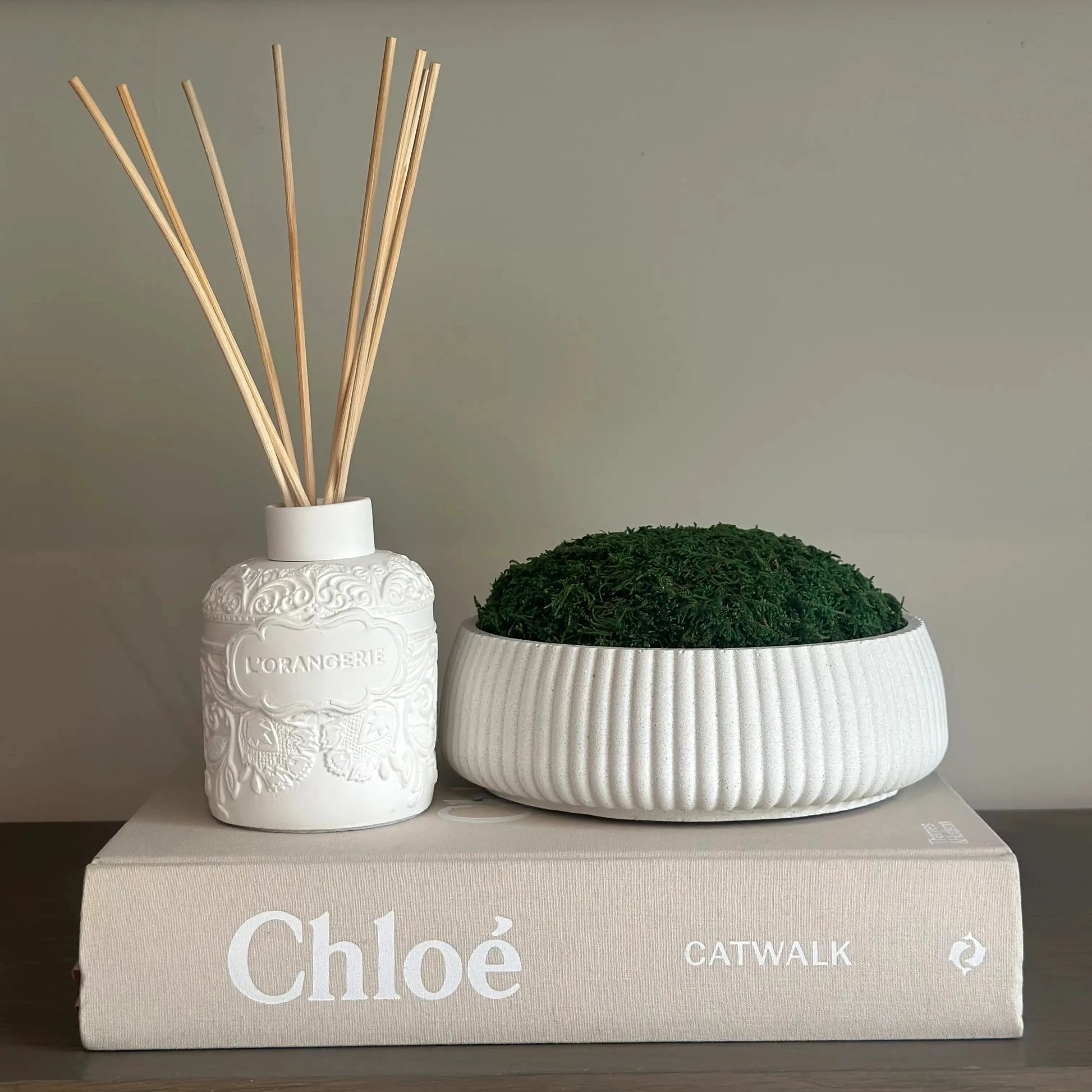 Chloe designer hardback coffee table book, displayed with white clay l'orangerie reed diffuser with beautiful spiced orang scent, and white handmade mineral bowl with preserved green natural moss. All available to purchase from Albury House Interiors, home decor and accessories in the UK. 