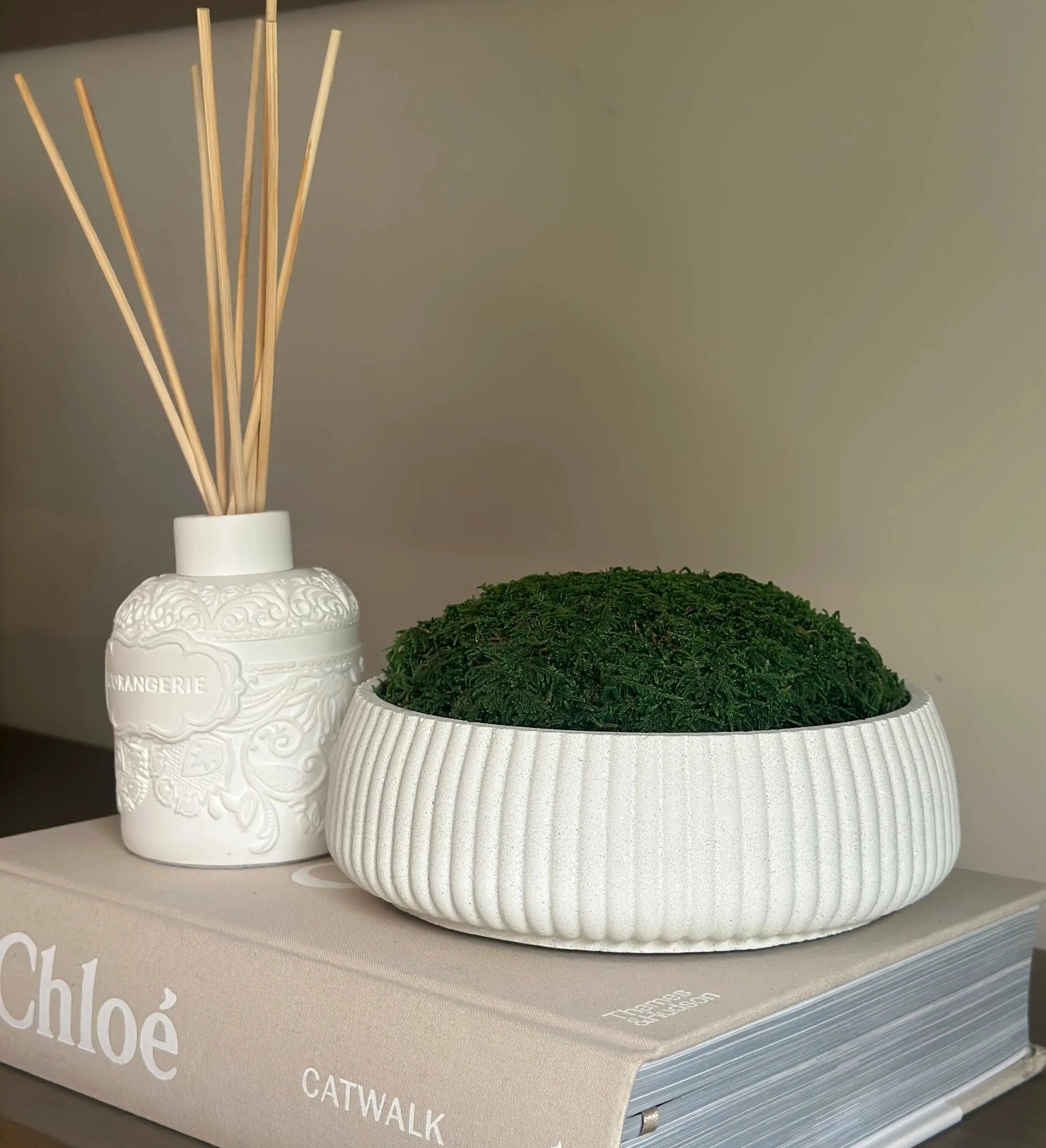 Chloe designer hardback coffee table book, displayed with white clay l'orangerie reed diffuser with beautiful spiced orang scent, and white handmade mineral bowl with preserved green natural moss. All available to purchase from Albury House Interiors, home decor and accessories in the UK. 