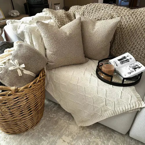 Neutral coloured soft throws and taupe boucle cushions on a bespoke sofa withCruz black round coffee table tray, displaying a Chanel designer book and a cup of tea. ,All available to purchase at Albury House Interiors. 