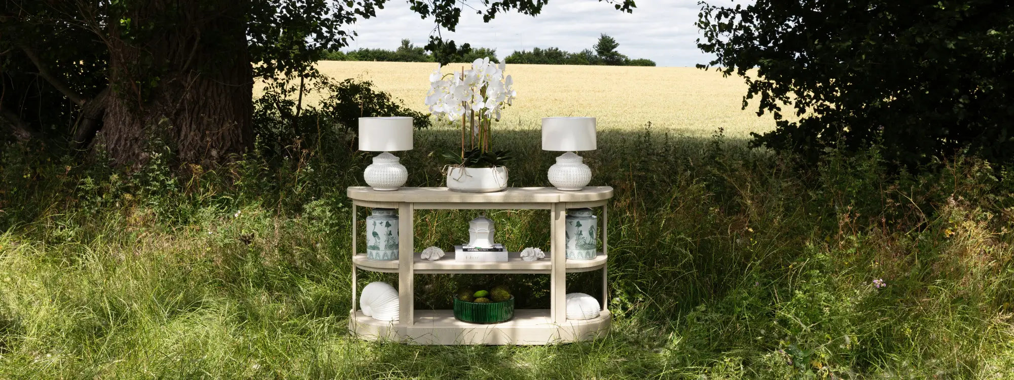 sancerre-limed-wooden-veneer-console-table-with-shelves-in-a-green-field-displaying-cream-ceramic-dimple-lamps-with-linen-shades-faux-potted-orchid-in-stone-pot-green-and-white-ostrich-ginger-jars-ceramic-snail-shell-and-faux-urchin-at-albury-house-interiors-homewares-store-essex
