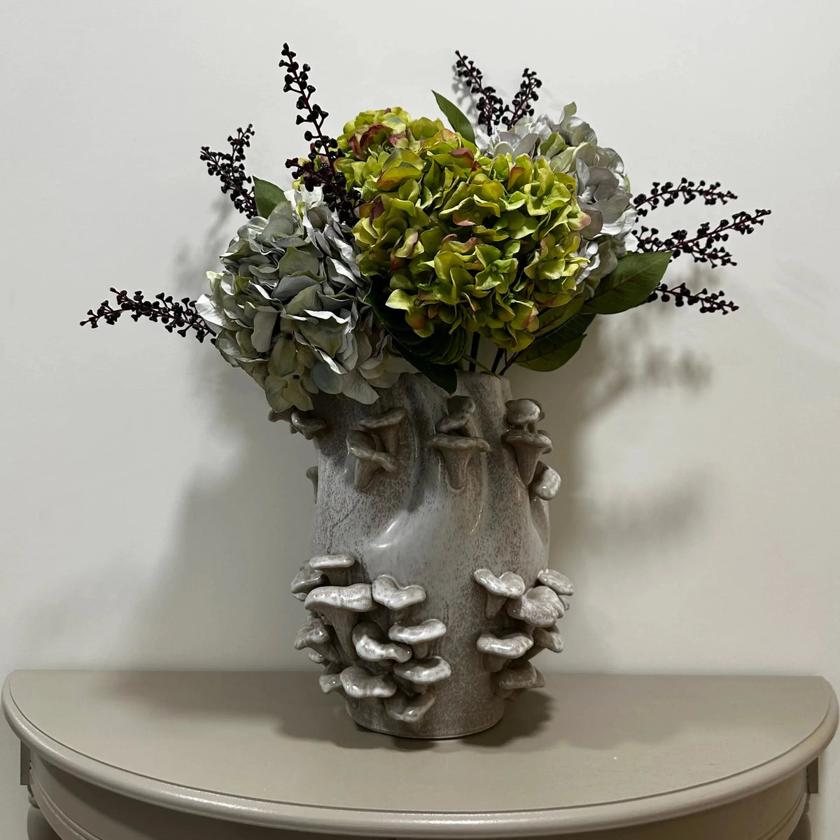 Grey speckled white ceramic vase with 3D fungi mushrooms and an artificial floral arrangement featuring green and silver hydrangea blooms and dark twig accents, displayed indoors on a half moon wooden table. Available to purchase at Albury House Interiors, Essex. 