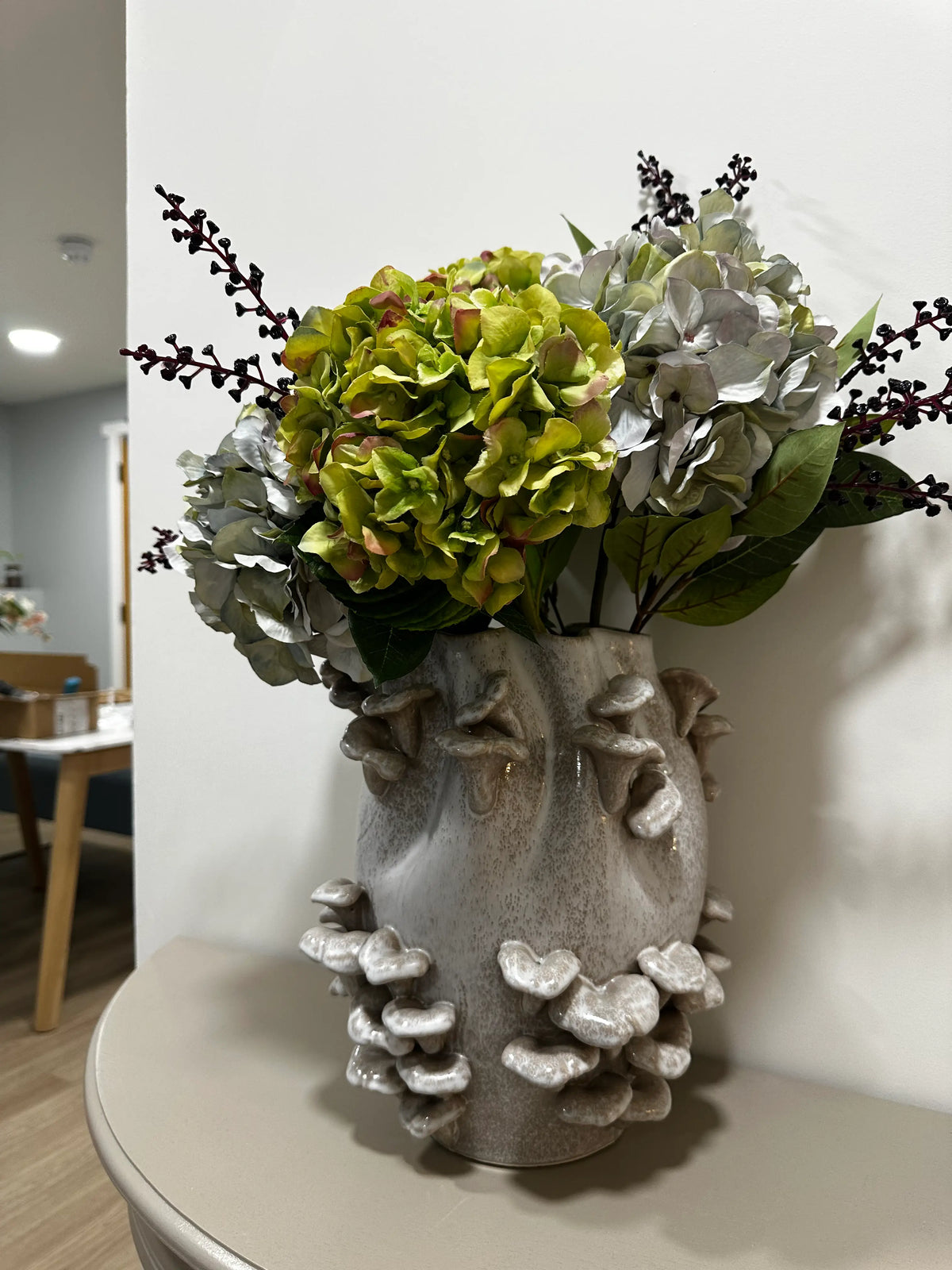 Grey speckled white ceramic vase with 3D fungi mushrooms and an artificial floral arrangement featuring green and silver hydrangea blooms and dark twig accents, displayed indoors on a half moon wooden table. Available to purchase at Albury House Interiors, Essex. 