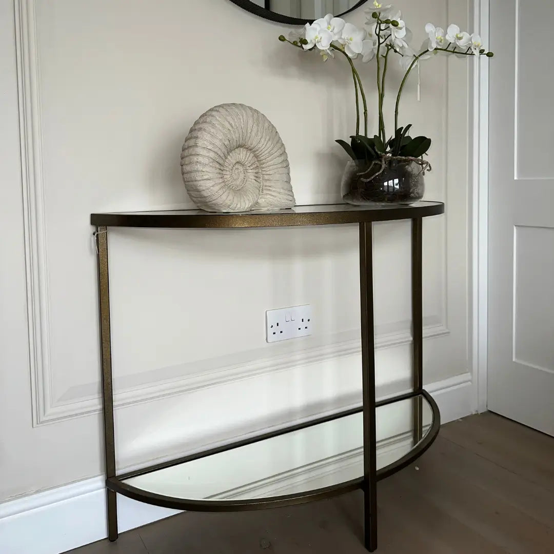 Half moon shaped console table with a bronze metal frame, a clear glass top, and a mirrored glass bottom shelf. Dsplayed with a faux fossil shell and faux orchid in glass pot. Available to purchase from Albury House Interiors.