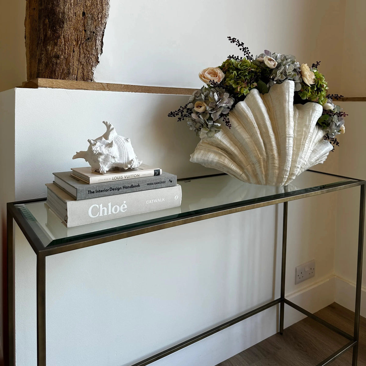 Antique bronze minimal console table with glass top, displaying our extra large clam shell with faux hydrangeas and peonies, and a stack of designer coffee table books with a faux white conch shell. All items available to purchase at Albury House Interiors, homewares and accessories in Matching Tye, Essex. 