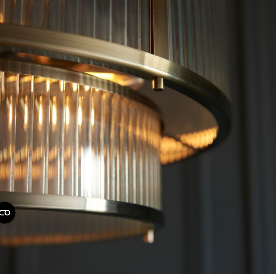 A two-tier drum pendant chandelier light with a brushed brass frame and clear ribbed glass panels, suspended from a matching chain against a white background, available from Albury House Interiors.