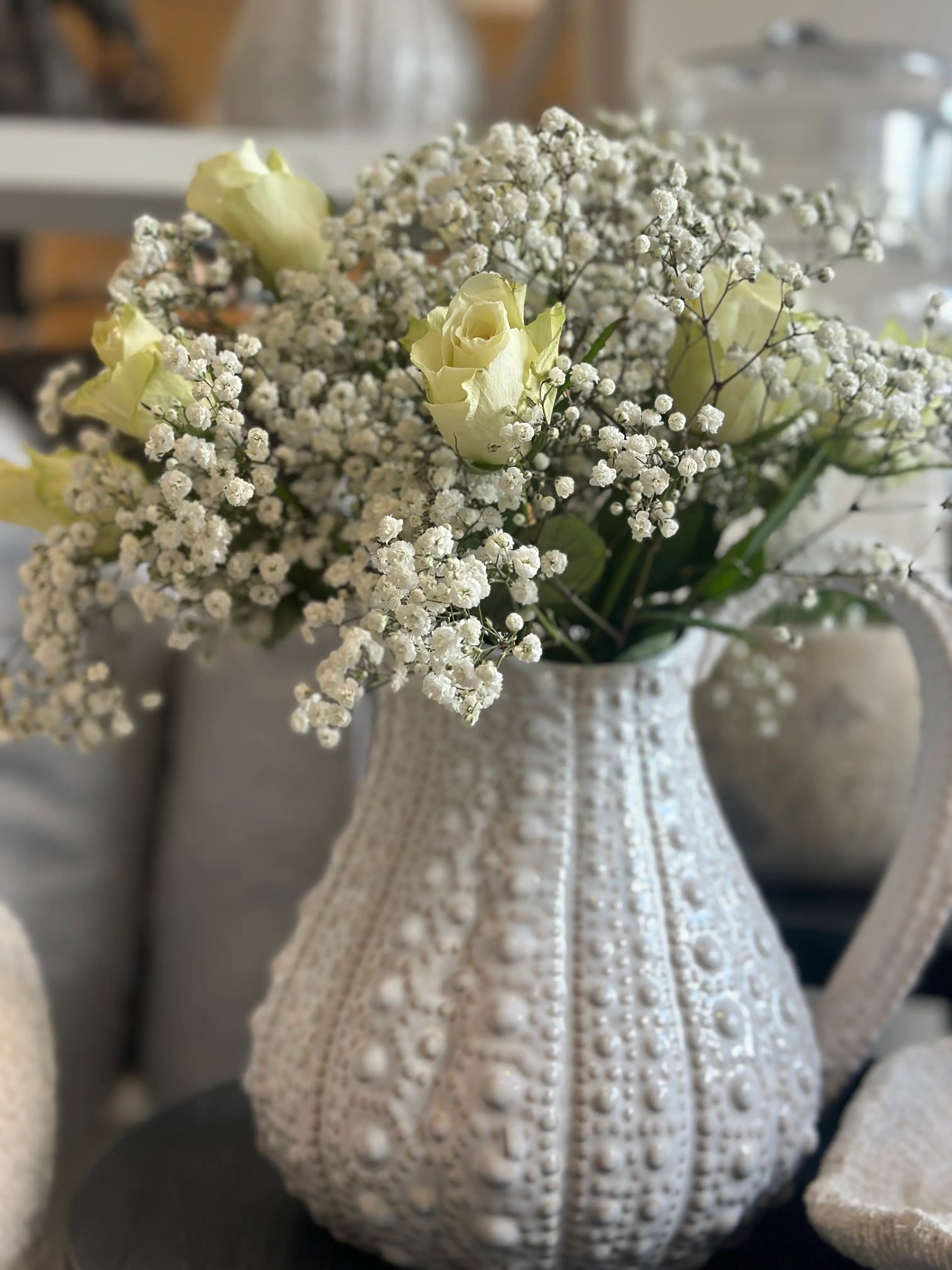White ceramic urchin jug vase, with dimpled all over texture, large handle and spout. Beautiful in all interiors and rooms due to its versatile colour and shape. Displayed with white roses and baby breath flowers. Available to purchase at Albury House Interiors, homewares and decor in Essex. 